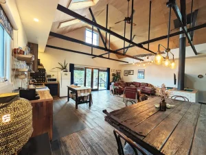 Spacious open-plan kitchen and living area with rustic wooden floors, exposed ceiling beams, large windows, a dining table, cozy sofas, and warm lighting, creating an inviting and stylish atmosphere.
