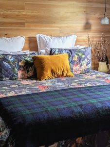 A cozy bed with floral bedding, three white pillows, and a mustard yellow pillow sits against a wooden wall. A blue plaid blanket is at the foot, and a vase with branches is on a nightstand beside a hanging lamp.
