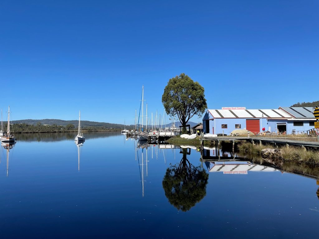 A calm river reflects sailboats, a tree, and a blue building with a red door under a clear blue sky. The water is still, creating a mirror-like effect with the boats and shoreline vegetation.