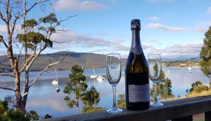 A bottle of sparkling wine and two filled glasses are set on a wooden railing overlooking a calm lake with sailboats, trees, and hills in the background under a partly cloudy sky.