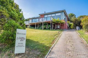 A modern two-story house with a brick and glass exterior sits on a grassy hill, accessed by a paved driveway. A sign out front reads The Beach House with contact details for inquiries. Shrubs and trees surround the property.