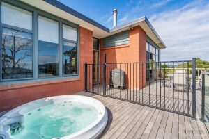 A modern house with large windows and a patio deck featuring a round outdoor hot tub, separated from the seating area by a black metal fence. Trees and blue sky are visible in the background.