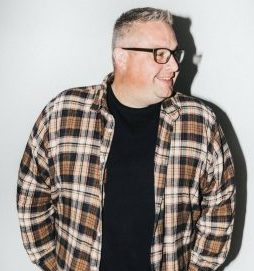 A man with short gray hair and glasses smiles while looking to his left. He wears a black shirt under a plaid button-up. The background is plain white, and a shadow is cast to his right.