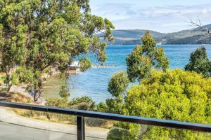 View from a balcony overlooking a blue lake or bay with a small dock, surrounded by green trees and rolling hills under a partly cloudy sky.