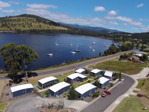 Aerial view of small white-roofed cabins near a road, beside a lake with boats, surrounded by green hills under a blue sky with scattered clouds.