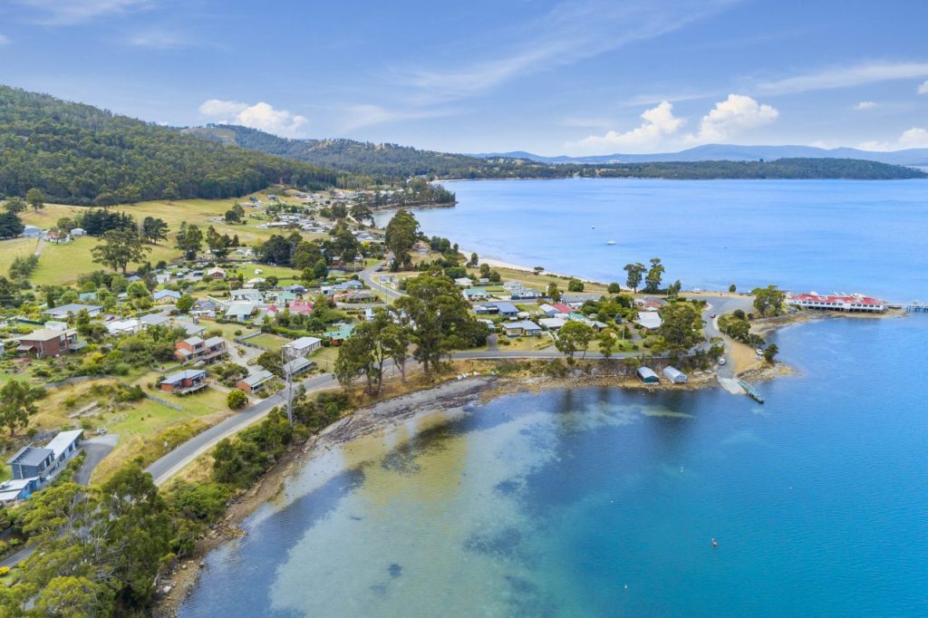 A coastal town with houses scattered near the shoreline, bordered by clear blue water and green hills, with a curving road along the bay and boats visible in the distance under a partly cloudy sky.