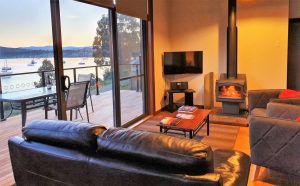 A cozy living room with a black leather couch, wood-burning stove, TV, and coffee table, featuring large windows overlooking a deck with patio furniture and a scenic lake with boats at sunset.