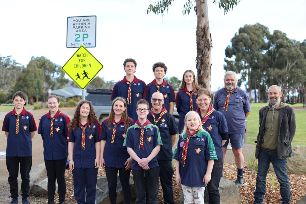 A group of Scouts and adult leaders in uniforms stand outdoors near a street with a “Watch for Children” sign and trees in the background, smiling and posing for the photo.