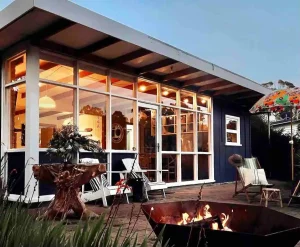 A cozy modern house with large windows is warmly lit inside. There are patio chairs, a colorful umbrella, and a fire pit with flames in the foreground, creating a welcoming outdoor space.