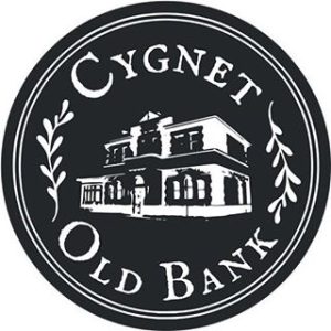 A circular black and white logo features an illustration of a historic building at its center, with the words Cygnet Old Bank written around the edge and decorative leaf accents on either side.