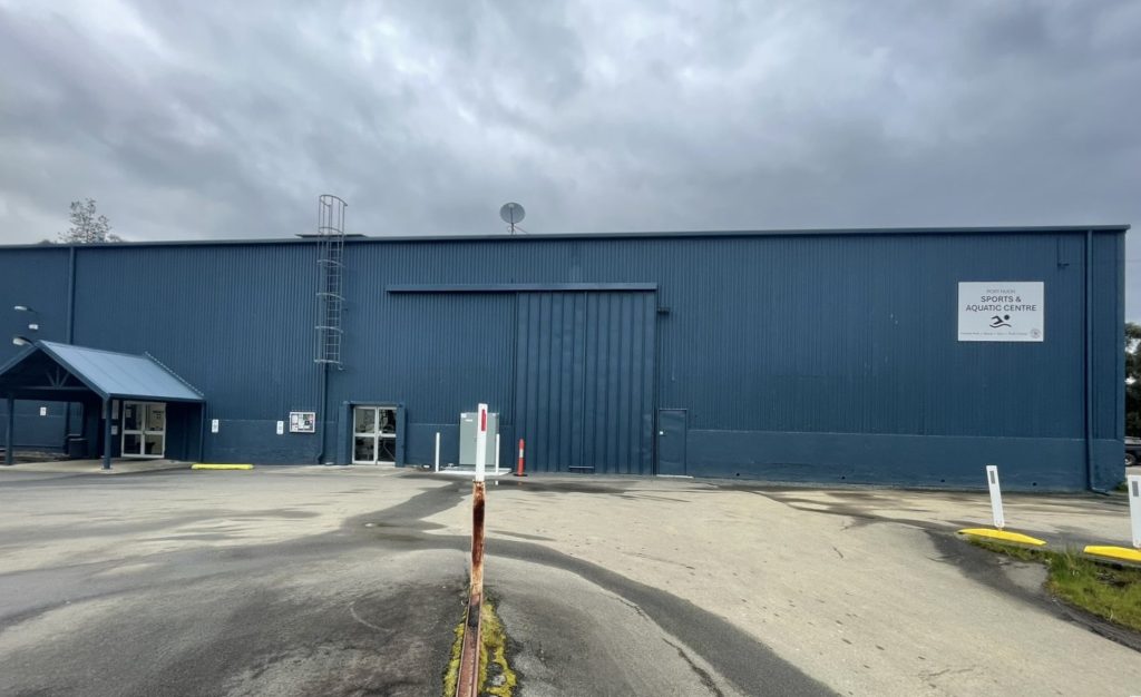 A large blue metal building with a small covered entrance on the left, a wide central garage door, and a sign reading “Sports Aquatic Centre” on the right under a cloudy sky. The ground is wet and mostly empty.
