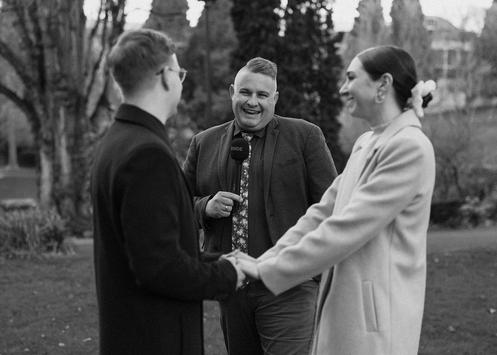 A smiling officiant holds a microphone while a couple stands facing each other, holding hands and smiling, outdoors in a park setting with trees in the background.