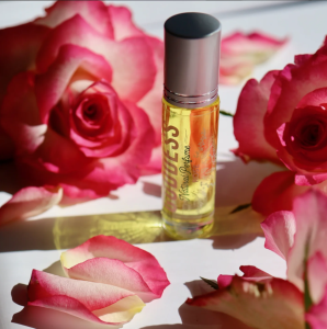 A glass perfume bottle with a silver cap is surrounded by pink and white roses and rose petals on a white surface, lit by natural sunlight.