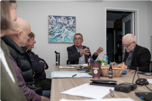 Five people sit around a meeting table in an office. One man gestures while speaking, others listen or take notes. Papers, notebooks, and drinks are on the table. A painting of ocean waves hangs on the wall behind them.
