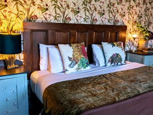 A cozy bed with white pillows and decorative animal-themed cushions, flanked by blue nightstands with lamps. The headboard is wooden, and the wall features floral wallpaper. The bed has a brown blanket at the foot.
