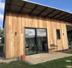 Small modern wooden house with a slanted metal roof, a sliding glass door, a small window, a black outdoor chair, a table, a potted plant, and a gravel area leading to a grassy lawn under a partly cloudy sky.