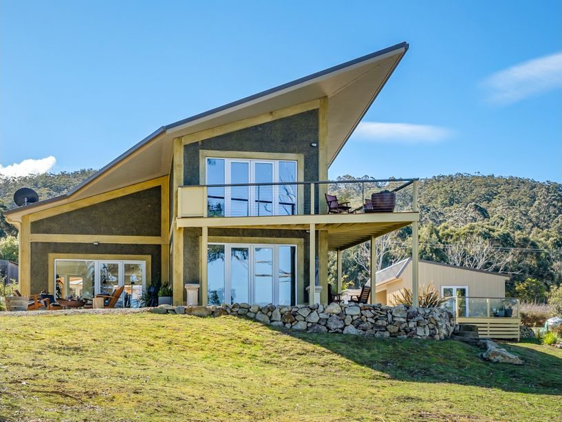 Modern two-story house with large windows, balcony, patio seating, and sloped roof, set on a grassy hill with mountains and trees in the background under a clear blue sky.