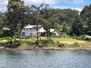 A large house with a light roof sits by the water, surrounded by trees and greenery. The scene is peaceful, with a grassy lawn leading down to the shoreline and a wooded area in the background.