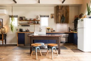 Cozy kitchen with rustic wooden floors, a central island with two wooden stools, blue lower cabinets, open shelves with dishes, hanging plants, and a white refrigerator. Warm, inviting atmosphere with natural light.