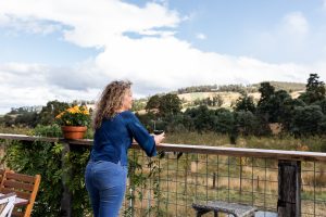 A woman with curly hair, wearing a blue shirt and jeans, stands on a wooden deck holding a cup, looking out at a scenic landscape of hills, trees, and a partly cloudy sky. A potted plant sits on the railing nearby.