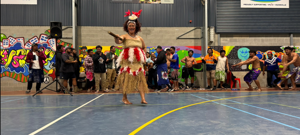 A performer in traditional Pacific Island attire dances in a gymnasium, while a group of people in colorful outfits watch from the background. The walls feature murals and company signs.