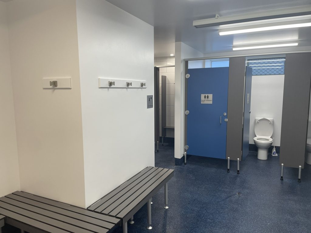 A clean public restroom with blue floors, gray benches, wall hooks, and several stalls, one with an open blue door revealing a toilet. Fluorescent lights brighten the space.