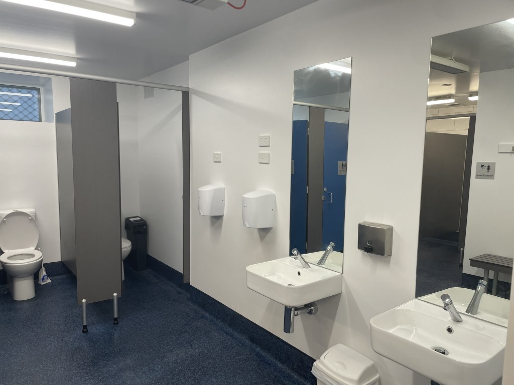 A clean public restroom with blue floors, white walls, two sinks with mirrors, soap dispensers, a toilet stall, and a bench in the corner. The space is brightly lit with overhead lights.