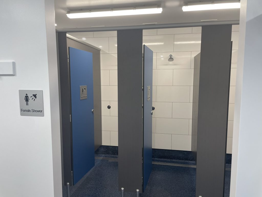 Three shower stalls with blue doors and gray partitions in a tiled bathroom. A sign on the wall to the left indicates this is the female shower area. The middle stall is open, revealing a showerhead inside.