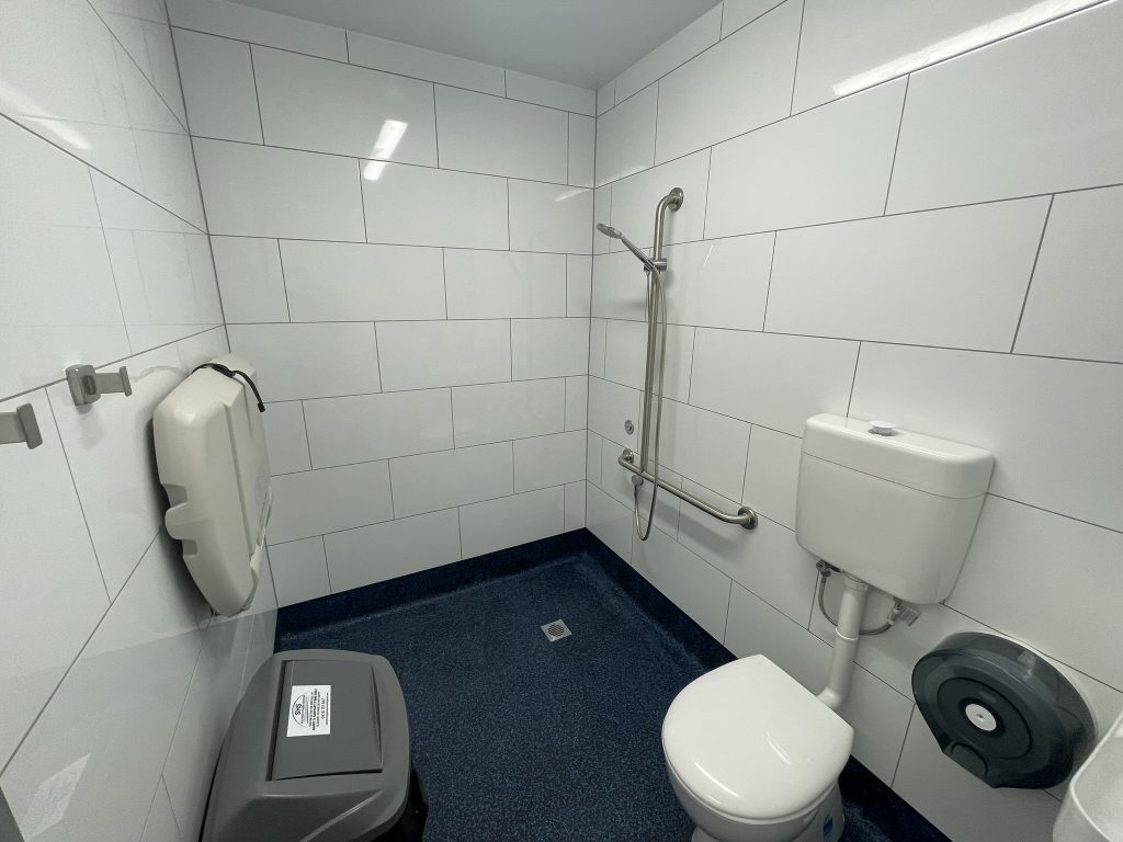 Accessible restroom with large white tiles, blue floor, toilet, toilet paper dispenser, wall-mounted trash bin, baby changing station, and metal grab bars on the wall for support.