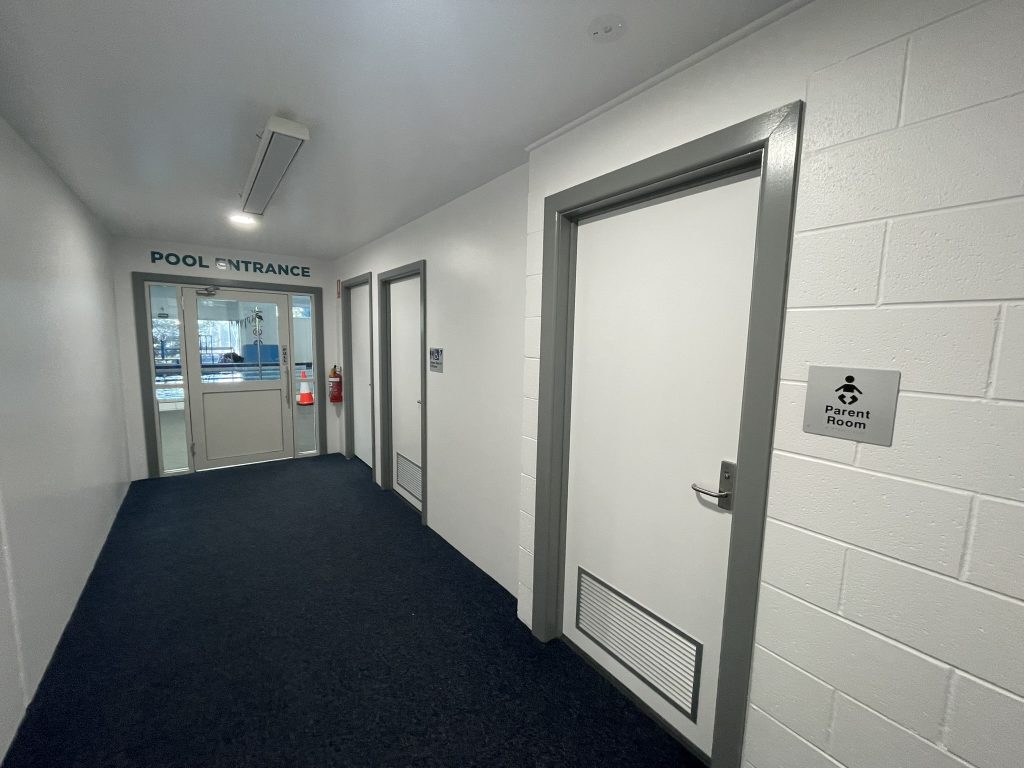 A hallway with white walls and blue carpet leads to a pool entrance with glass doors. To the right, there is a door labeled Parent Room with a sign showing a parent and child icon.
