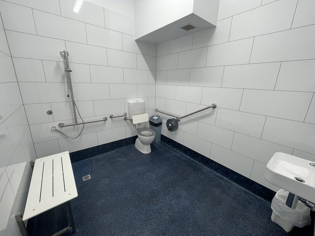 Accessible bathroom with shower, toilet, sink, grab bars, and a fold-down seat on dark blue speckled flooring.