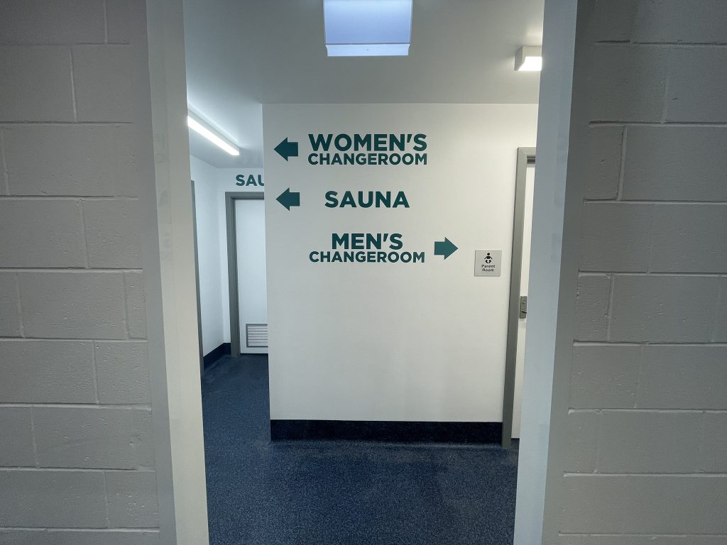 A hallway with directional signs on a white wall indicating left for “Women’s Changeroom,” straight ahead for “Sauna,” and right for “Men’s Changeroom.” The floor and lower walls are blue.