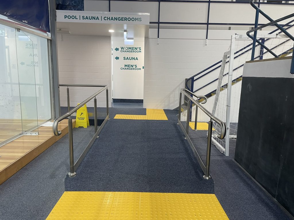 An indoor pool facility with accessible ramps and yellow tactile paving leading to womens and mens changerooms and a sauna; signs are visible above the doors and a wet floor caution sign is on the left.