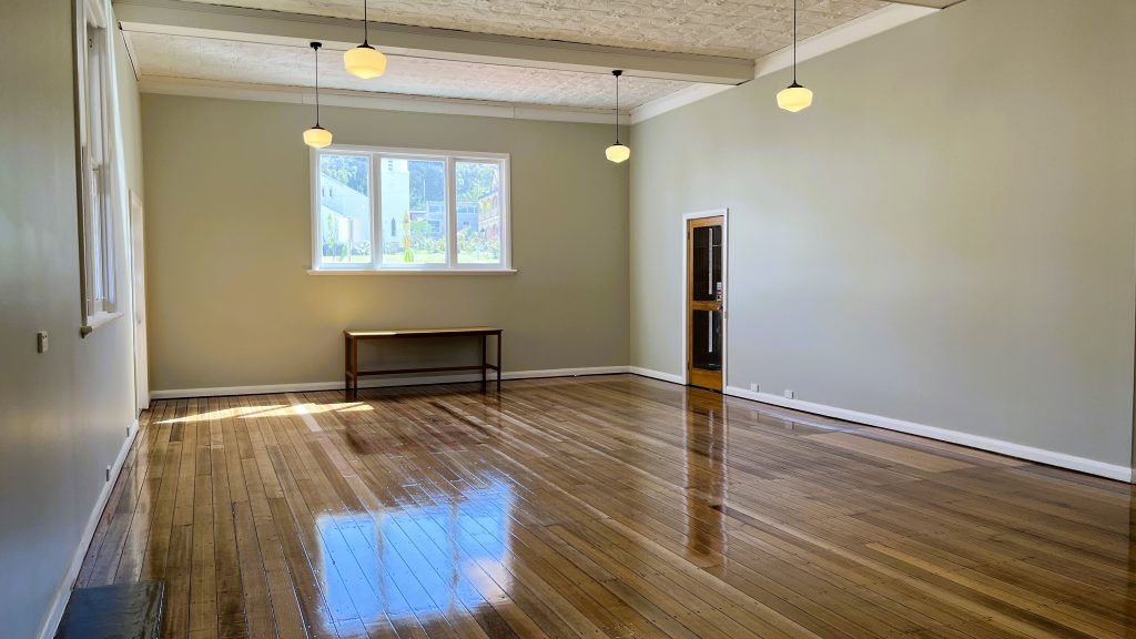 A spacious, empty room with polished wooden floors, light gray walls, five hanging pendant lights, large windows letting in natural light, and a wooden table against the back wall under the windows.