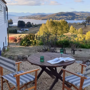 A round wooden table with two striped chairs sits on a gravel patio, overlooking a scenic landscape of hills, trees, and a lake with mountains in the distance. An open book and two green glasses are on the table.
