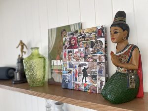 A wooden shelf displays a decorative green glass bottle, a book, a collage photo frame, a small statuette, and a colorful figurine of a woman in traditional attire with hands in a prayer gesture.