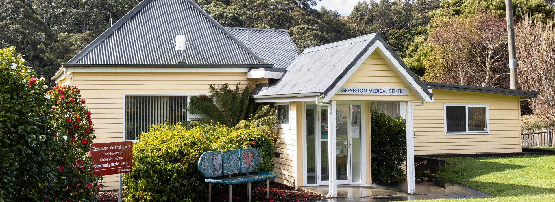 A small, single-story yellow building with a metal roof and a sign reading Geeveston Medical Centre stands surrounded by greenery, trees, and a garden bench near the entrance on a clear day.