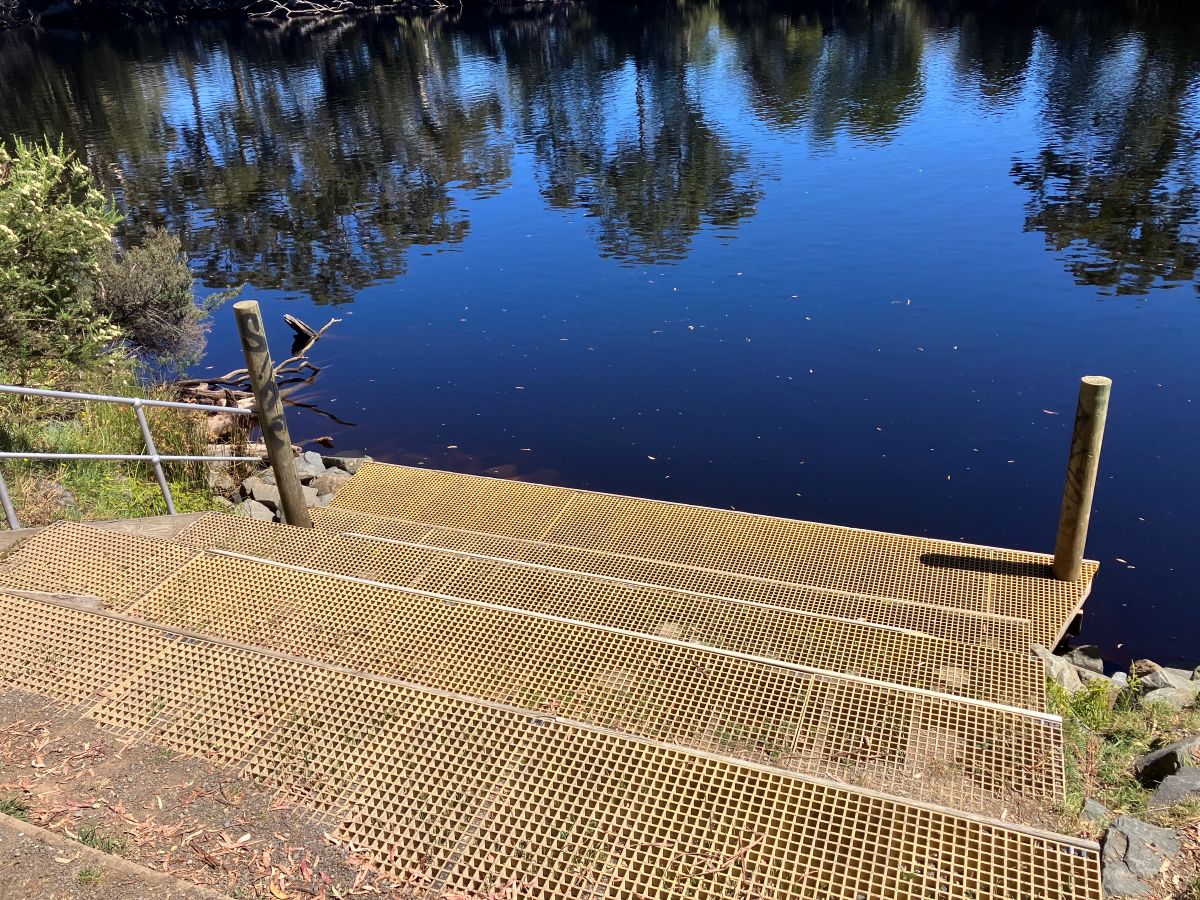 Metal steps lead down to a calm, blue river surrounded by trees, with reflections of the sky and foliage visible on the water. Wooden and metal railings border the steps.