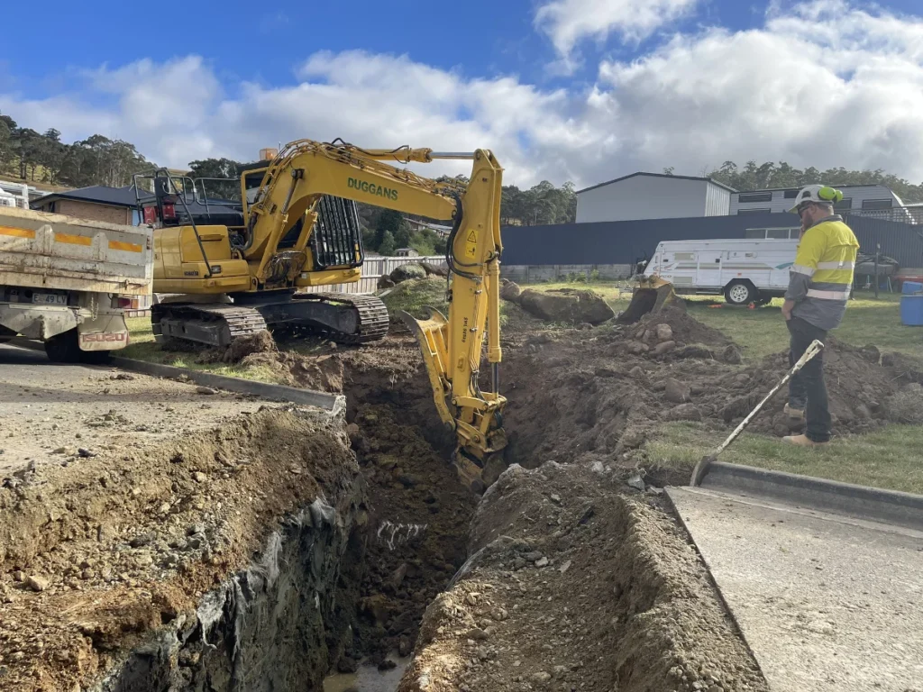 A construction worker stands near a deep trench while a yellow excavator digs dirt. There is a truck and trailer in the background on a partly cloudy day at a construction site near houses.