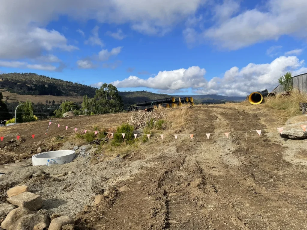 Dirt construction site with tire tracks, scattered rocks, a concrete ring structure, and large yellow pipes. Red and white bunting lines part of the area, with hills, trees, and a cloudy blue sky in the background.