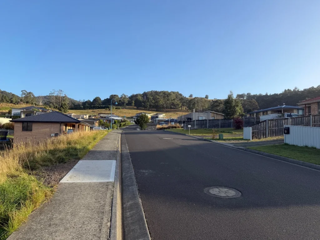 A quiet suburban street with single-story houses, grassy lawns, and a clear blue sky. Hills with trees are visible in the background, and the road is empty with no vehicles or people present.