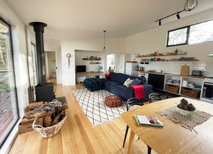 Bright, open-plan living space with a navy sofa, patterned rug, wood stove, firewood basket, dining table, and modern kitchen. Large windows let in natural light, and shelves hold various kitchen items and decor.