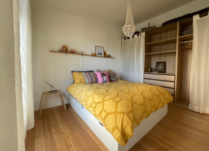 A cozy bedroom with a yellow bedspread, colorful pillows, a wooden floor, open closet with shelves, and a small side table. A shelf with decor and framed art hangs above the bed, and a chandelier light fixture is overhead.