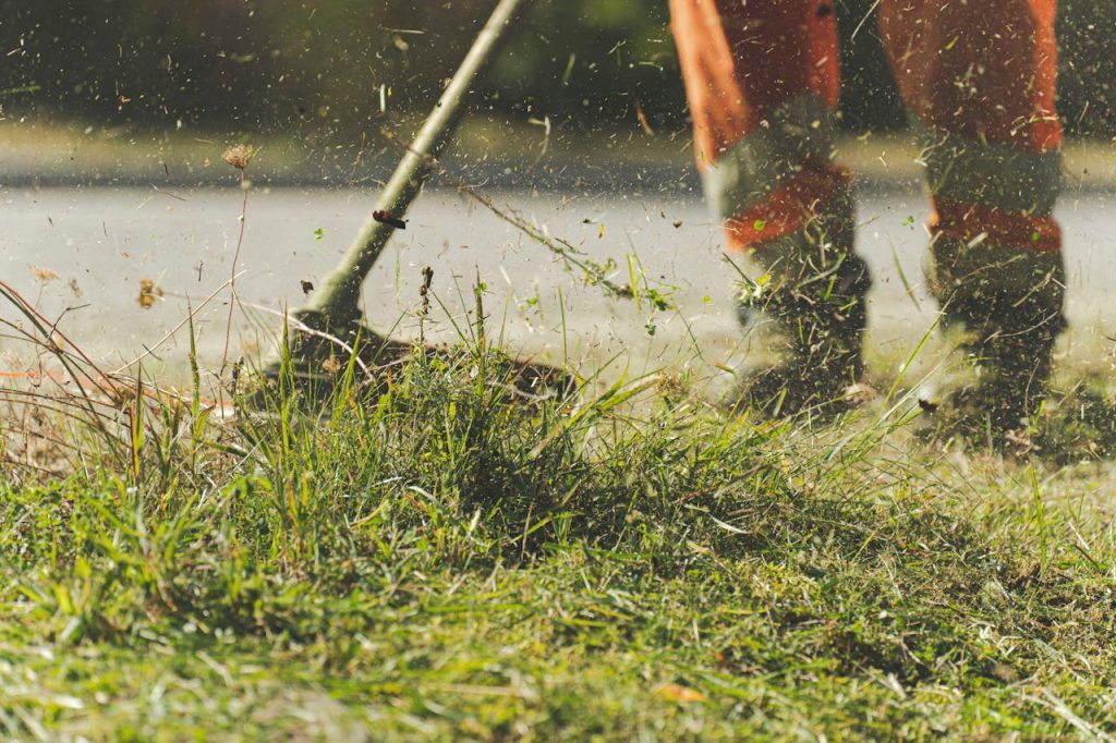 Close-up of a person wearing orange pants using a string trimmer to cut grass, with blades and debris flying in the air. The persons upper body is not visible.