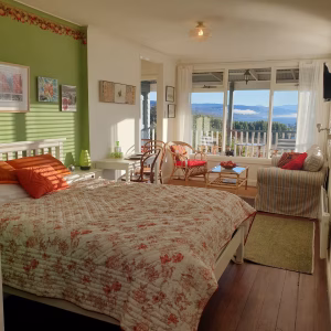 Bright, cozy bedroom with a floral quilt, orange and green accents, wooden floor, wicker chairs, a striped sofa, and large windows offering a scenic view of trees and mountains under a blue sky.