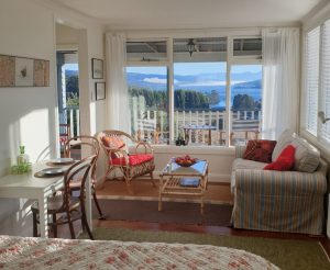 A bright, cozy sunroom with wicker furniture, a striped sofa, a small dining table, and large windows offering a scenic view of a lake, forest, and distant mountains. Sunlight fills the space.