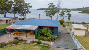 Blue house with a covered porch, surrounded by gardens, overlooking a bay and trees. A separate small building sits nearby, with water and forested hills visible in the background.