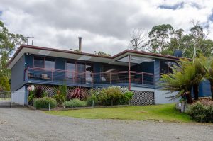 Modern single-story house with blue exterior, large glass windows, red railings, and a covered balcony. Surrounded by greenery, including shrubs and trees, with a gravel driveway in front and a partly cloudy sky overhead.