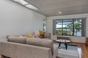 A cozy living room with two beige sofas, a small round table, and throw pillows. A large window reveals a scenic view of water and trees outside. The room has wooden flooring and a striped ceiling.