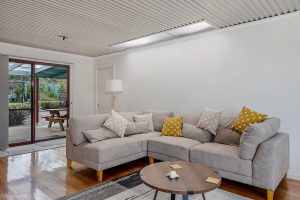 A modern living room with a large gray sectional sofa decorated with yellow and patterned throw pillows, a round wooden coffee table, and a floor lamp. Sliding glass doors open to a patio with outdoor seating and greenery.